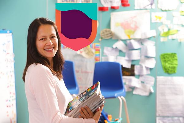 Teacher in a classroom, smiling and holding a pile of books